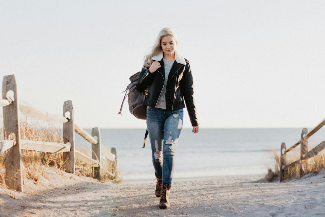 A women wearing a black jacket with jeans walking in the beach with a bag in hand